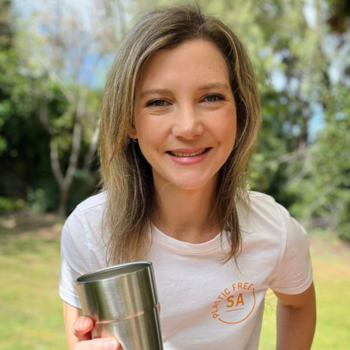 Smiling person outdoors holding a reusable metal cup, wearing a white T-shirt.